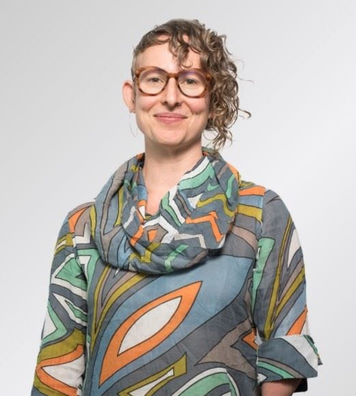 Lezlie looks at us full front with curls down one size of her head and her long sleeve rolled up just on that side of a cowl neck wild abstract print shirt. Her smiling cheeks greet us