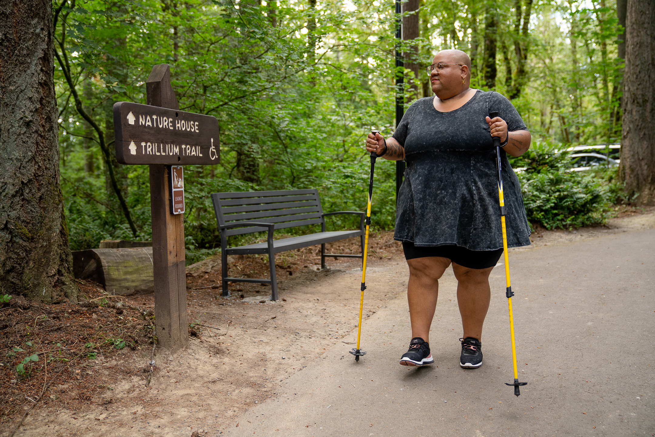 :img A disabled Black non-binary hiker uses trekking poles and looks at a wooden sign with “Nature House” and “Trillium Trail ♿” marked ahead. The shot is framed with the hiker walking toward the camera and the hiker sports a shaved head, glasses, a gray peplum shirt, black shorts, and black tennis shoes.